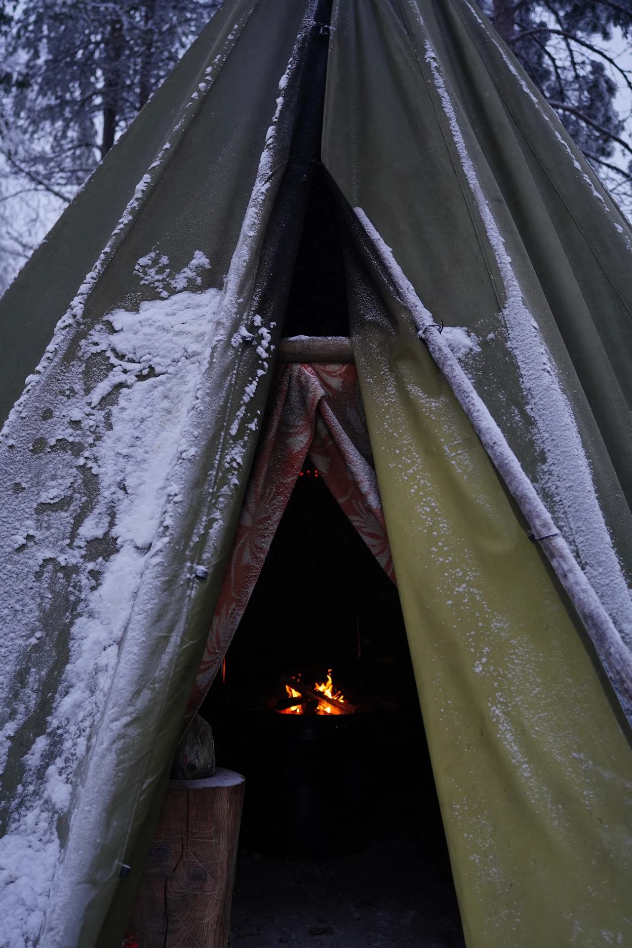 Snow-covered canvas hut with a small warming fire visible inside