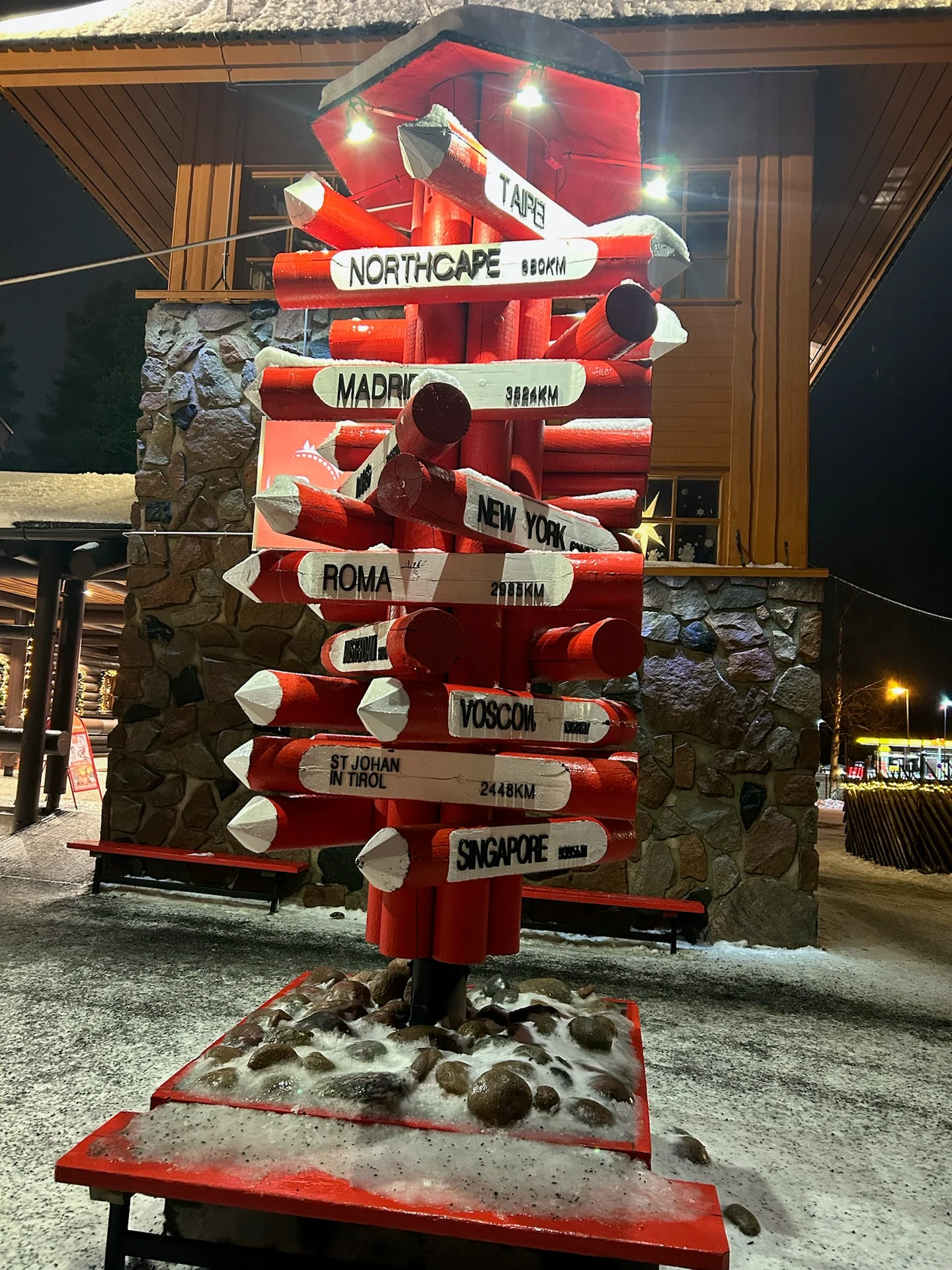 Red and white distance sign pointing from Santa Claus Village to cities around the world